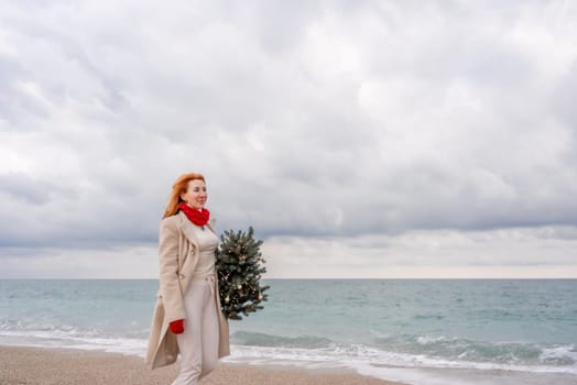 Redhead woman Christmas tree sea. Christmas portrait of a happy redhead woman walking along the beach and holding a Christmas tree in her hands. Dressed in a light coat, white suit and red mittens