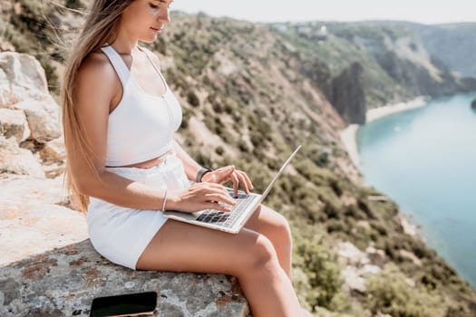 Digital nomad, Business woman working on laptop by the sea. Pretty lady typing on computer by the sea at sunset, makes a business transaction online from a distance. Freelance remote work on vacation