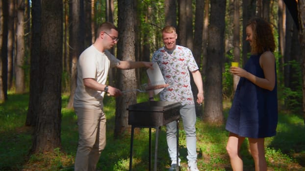 Man blows up coals on grill. Stock footage. Friends are standing at grill and blowing up coals in forest. Friends cook on grill and blow up coals for roasting meat outdoors in summer.