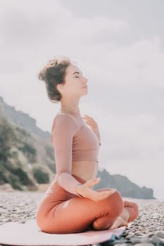 Young woman with long hair in white swimsuit and boho style braclets practicing outdoors on yoga mat by the sea on a sunset. Women's yoga fitness routine. Healthy lifestyle, harmony and meditation