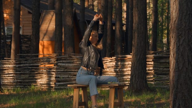 Beautiful young woman is sitting on bench and warming up. Stock footage. Young woman stretches out on bench waiting for date. Attractive woman stretches while sitting on bench in woods.