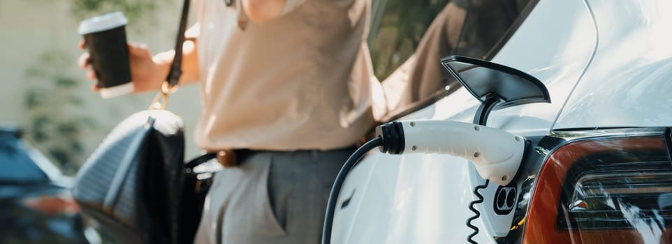 Young man with coffee cup, recharge electric car's battery from EV charging station in city commercial parking lot. Eco friendly EV car with urban and shopping lifestyle. Panorama Expedient