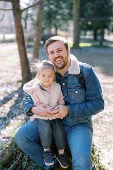 Happy dad with a little girl on his knees sit on a stump in a spring park. High quality photo