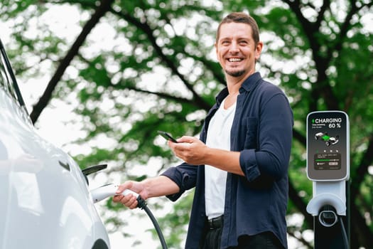 Man using smartphone online banking application to pay for electric car battery charging from EV charging station during vacation holiday road trip at national park or summer forest. Exalt