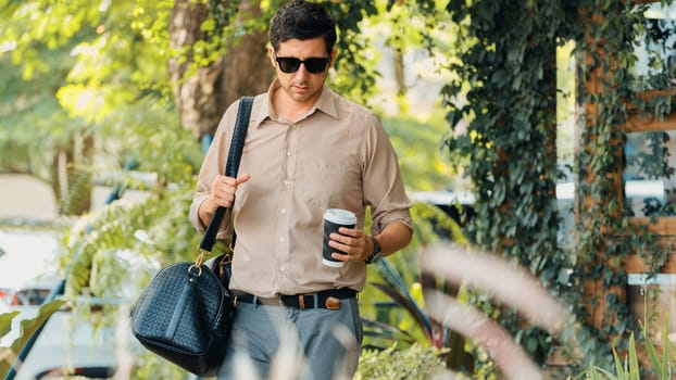 Young handsome man walk out of coffee shop with cafe garden, holding a coffee cup and a shopping bag. Modern happy carefree with sunglasses lifestyle. Expedient