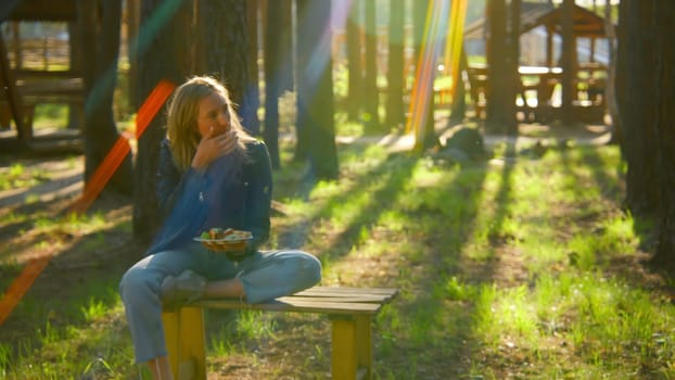 Young woman is sitting on bench and eating barbecue. Stock footage. Beautiful woman is sitting alone and eating barbecue in park in summer. Woman with barbecue plate in nature on sunny summer day.