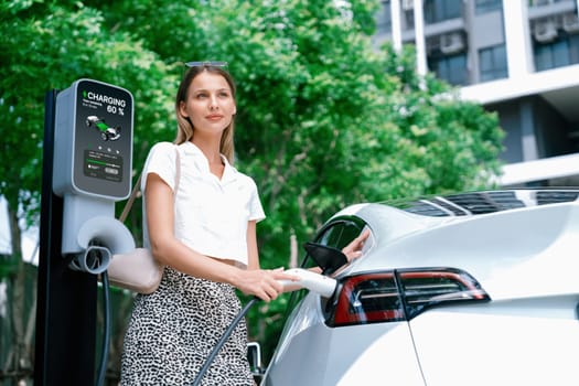 Young woman travel with EV electric car charging in green sustainable city outdoor garden in summer. Urban sustainability lifestyle by green clean rechargeable energy of electric BEV vehicle innards