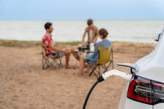 Alternative family vacation trip traveling by the beach with electric car recharging battery from EV charging station with blurred family enjoying the seascape background. Perpetual