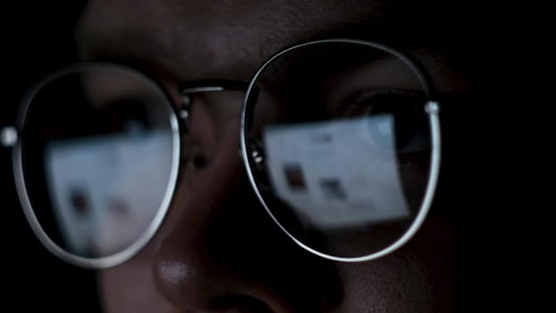 Man works on internet with the reflection of a monitor inside glasses. Close up of male face wearing glasses with the screen reflected in lenses isolated on black background.