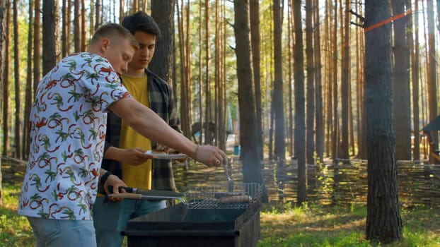Men put barbecue on plate in nature. Stock footage. Men cooked sausages on grill in forest on sunny summer day. Men put barbecue meat on plate in nature.