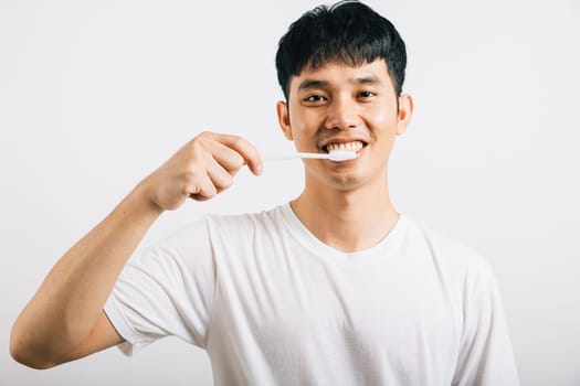 Portrait of a smiling Thai young man confidently brushing his teeth for oral health. Studio shot isolated on white background, promoting dental and oral care.