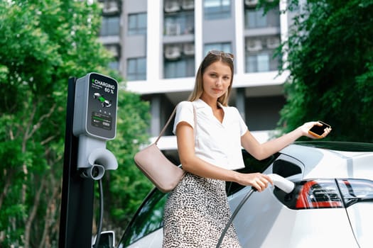 Young woman travel with EV electric car charging in green sustainable city outdoor garden in summer. Urban sustainability lifestyle by green clean rechargeable energy of electric BEV vehicle innards