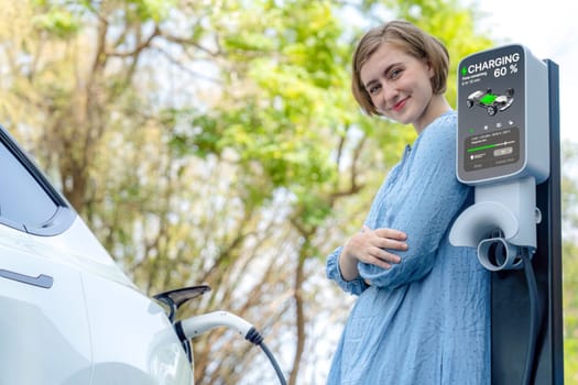 Holiday road trip vacation by the green countryside nature with beautiful young woman recharging electric vehicle with alternative energy. Environmental friendly travel wit EV car. Perpetual