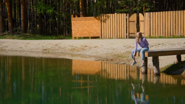 Young woman is sitting on edge of pier alone. Stock footage. Beautiful young woman throws stones into water on wooden pier. Young woman is sitting on pier and throwing stones on sunny summer day.