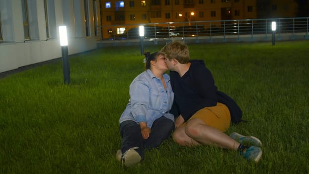 Young couple kissing on lawn. Media. Couple in love kissing sitting on green grass in park at night. Night park with kissing happy couple of overweight people.