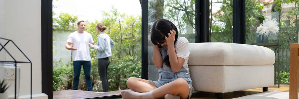 Stressed and unhappy young girl huddle in corner, cover her ears blocking sound of her parent arguing in background. Domestic violence at home and traumatic childhood depression. Panorama Synchronos
