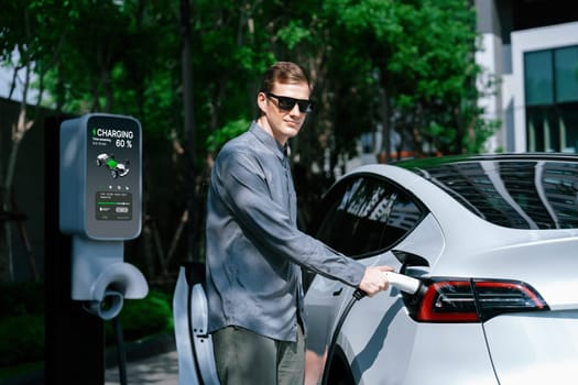 Young man travel with EV electric car charging in green sustainable city outdoor garden in summer shows urban sustainability lifestyle by green clean rechargeable energy of electric vehicle innards