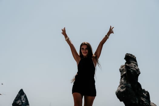 Woman travel sea. Young Happy woman in a long red dress posing on a beach near the sea on background of volcanic rocks, like in Iceland, sharing travel adventure journey