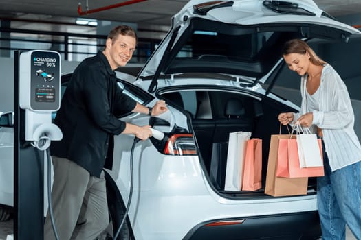 Young couple travel with EV electric car to shopping center parking lot charging in downtown city showing urban sustainability lifestyle by green clean rechargeable energy of electric vehicle innards