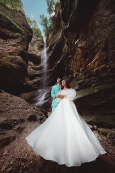 Wedding of a beautiful couple, the bride is dressed in a white dress. Stylish shooting against the backdrop of a natural landscape with a waterfall in the mountains in summer.