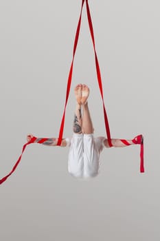 Sportsman performance on a red canvases. Bald guy in a light sport suit is doing an acrobatic elements hanging on a rope in a studio isolated on white background. Dancing in the air with balance.