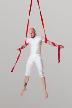 Sportsman performance on a red canvases. Bald male in a light sport suit is doing an acrobatic elements hanging on a rope and looking down in a studio isolated on white background. Dancing in the air with balance.