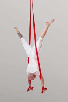 Sportsman performance on a red canvases. Bald man in a light sport suit is doing an acrobatic elements hanging on a rope upside down in a studio isolated on white background. Dancing in the air with balance.
