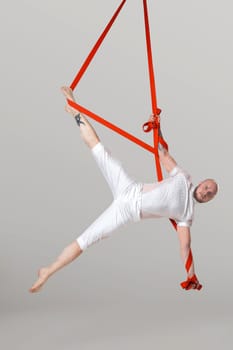 Sportsman performance on a red canvases. Bearded male in a light sport suit is doing an acrobatic elements hanging on a rope in a studio isolated on white background. Dancing in the air with balance.