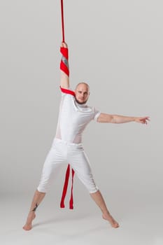 Sportsman performance on a red canvases. Bearded man in a light sport suit is doing an acrobatic elements hanging on a rope and looking at the camera in a studio isolated on white background. Dancing in the air with balance.