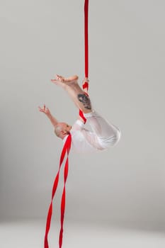 Gymnast performance on a red canvases. Athletic guy in a light sport suit is doing an acrobatic elements hanging on a rope in a studio isolated on white background. Dancing in the air with balance.