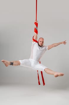 Gymnast performance on a red canvases. Athletic man in a light sport suit is doing an acrobatic elements hanging on a rope in a studio isolated on white background. Dancing in the air with balance.
