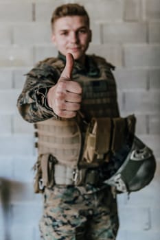 The soldier makes a gesture of success with his hand. A soldier in full war gear stands in front of a stone wall and shows the ok sign with his finger.