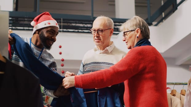 Retail assistant in xmas adorn fashion boutique helping old couple during their Christmas frenzy shopping spree. Worker offering advice to aged clients during festive holiday season