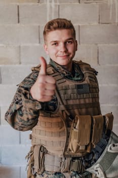 The soldier makes a gesture of success with his hand. A soldier in full war gear stands in front of a stone wall and shows the ok sign with his finger.