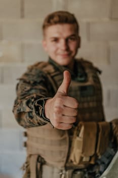 The soldier makes a gesture of success with his hand. A soldier in full war gear stands in front of a stone wall and shows the ok sign with his finger.