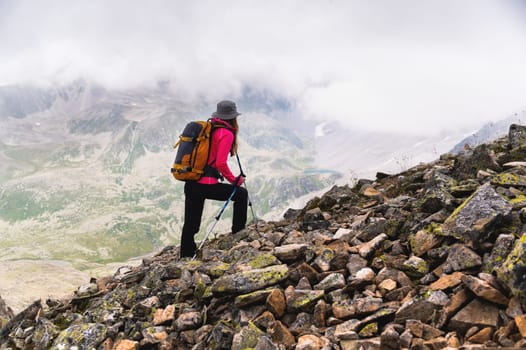 A woman traveler goes to the top of the mountain. A girl with trekking poles and a backpack stopped to admire the fog approaching her in the mountains.