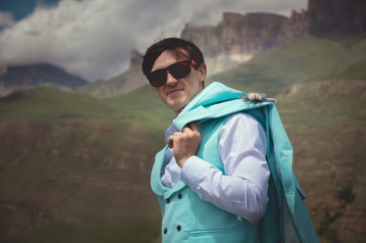 Portrait of a cheerful Caucasian guy in black glasses, holding his jacket over his shoulder and looking down at the camera, against the backdrop of rocky mountains and low clouds. confident young man.