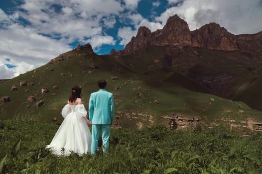 Wedding. A guy and a girl in a white dress stand on a green field opposite the mountains and look at the high rocky ridges.