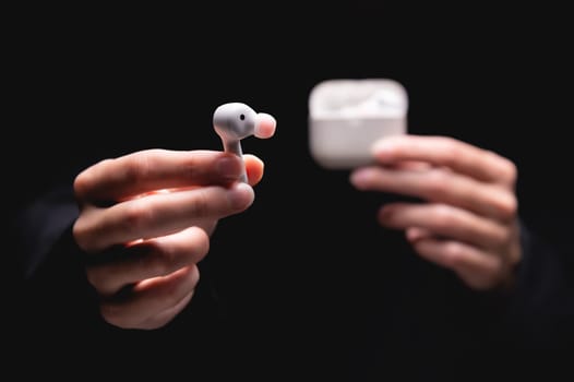 close-up of a woman taking a white wireless earphone out of the charger. Female hands touch the headphones of a portable gadget and present it forward to the camera on a black background.