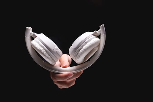 A hand holds headphones on a black background. Modern white headphones