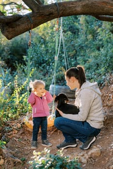 Little smiling girl stands near a swing and watches her mom petting a puppy on her lap. High quality photo