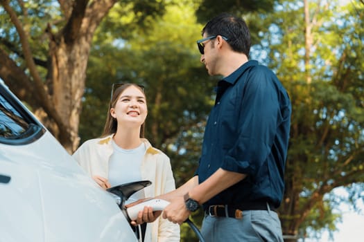 Young couple recharge electric car's battery from charging station in outdoor green city park in springtime. Rechargeable EV car for sustainable environmental friendly urban travel lifestyle.Expedient