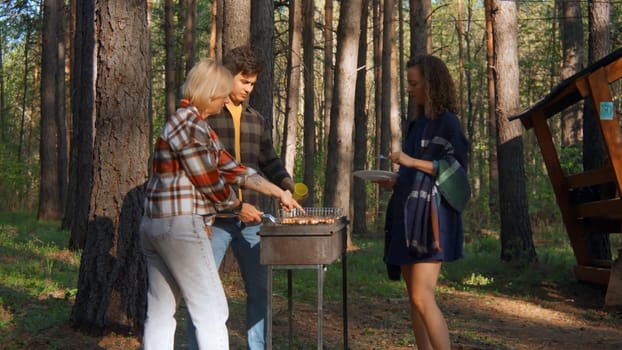 Friends are standing at barbecue in nature on sunny day. Stock footage. Group of friends in nature cook meat on grill. Friends cook meat on barbecue in sunny summer forest.