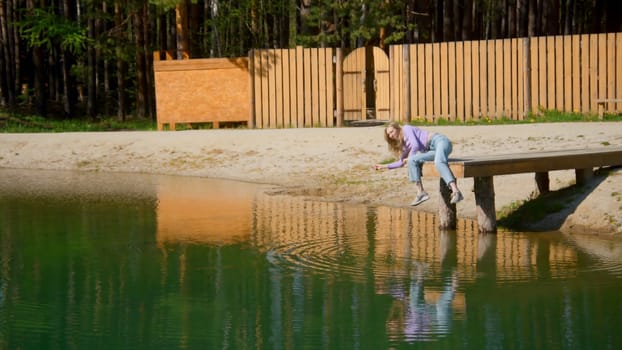 Young woman is sitting on edge of pier alone. Stock footage. Beautiful young woman throws stones into water on wooden pier. Young woman is sitting on pier and throwing stones on sunny summer day.