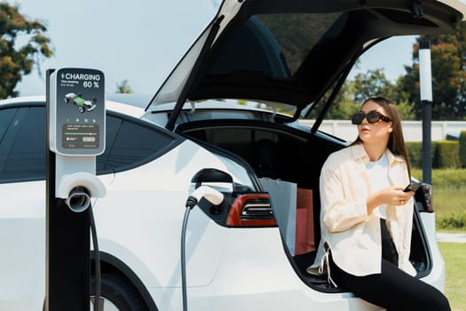 Young woman use smartphone to pay for electricity at public EV car charging station green city park. Modern environmental and sustainable urban lifestyle with EV vehicle. Expedient