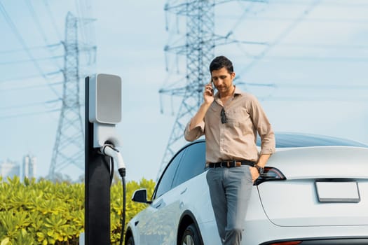 Man talking on the phone while recharge EV car battery at charging station connected to power grid tower electrical as electrical industry for eco friendly car utilization.Expedient