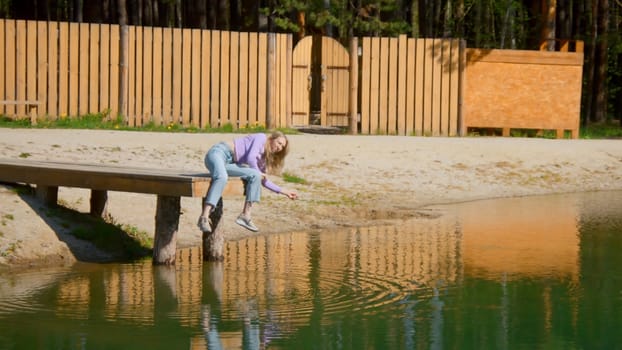 Young woman is sitting on edge of pier alone. Stock footage. Beautiful young woman throws stones into water on wooden pier. Young woman is sitting on pier and throwing stones on sunny summer day.