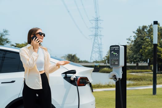 Young woman recharging EV car battery while talk on phone at charging station connected to electrical power grid tower facility as electrical industry for eco friendly vehicle utilization. Expedient