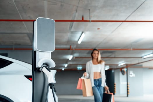 Young woman travel with EV electric car to shopping center parking lot charging in downtown city showing urban sustainability lifestyle by green clean rechargeable energy of electric vehicle innards