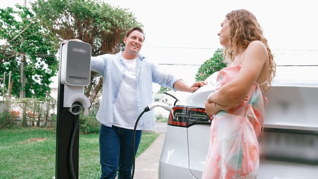 Happy and lovely couple with eco-friendly conscious recharging electric vehicle from EV charging station. EV car technology utilized as alternative transportation for future sustainability. Synchronos